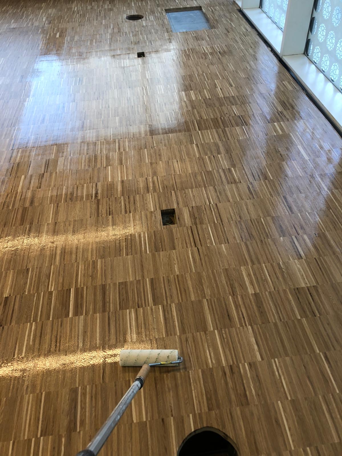 Close-up of timber block flooring being coated with a roller in a large interior, showing the contrast between finished and unfinished surfaces.