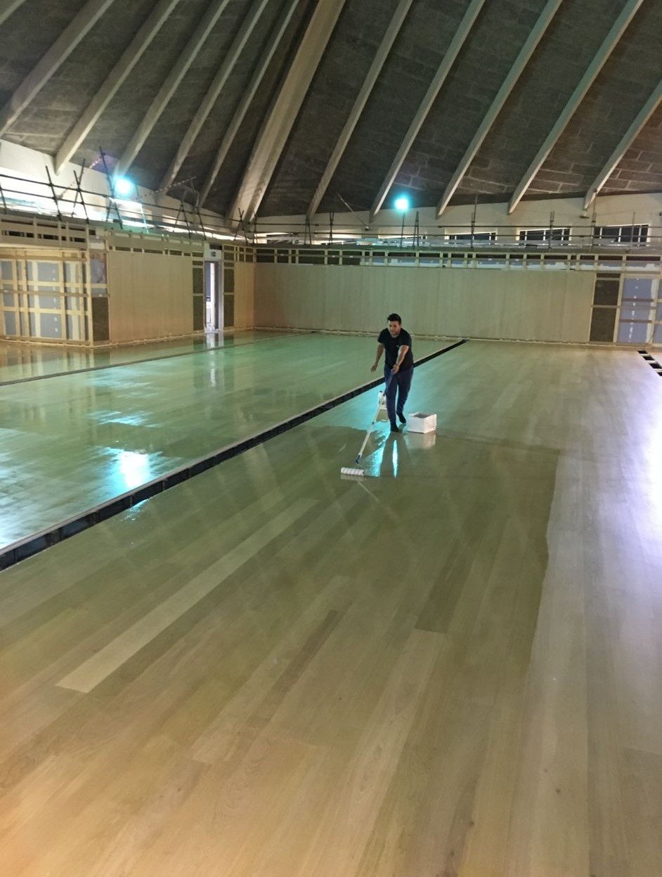 Flooring specialist applying a finishing coat with a roller across a large natural timber floor in a high-ceilinged architectural interior.