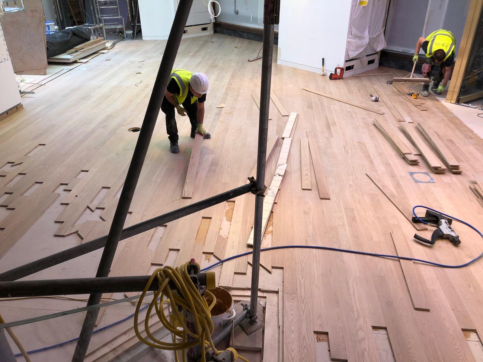 Two installers fitting long timber flooring planks in a large interior while construction works continue.