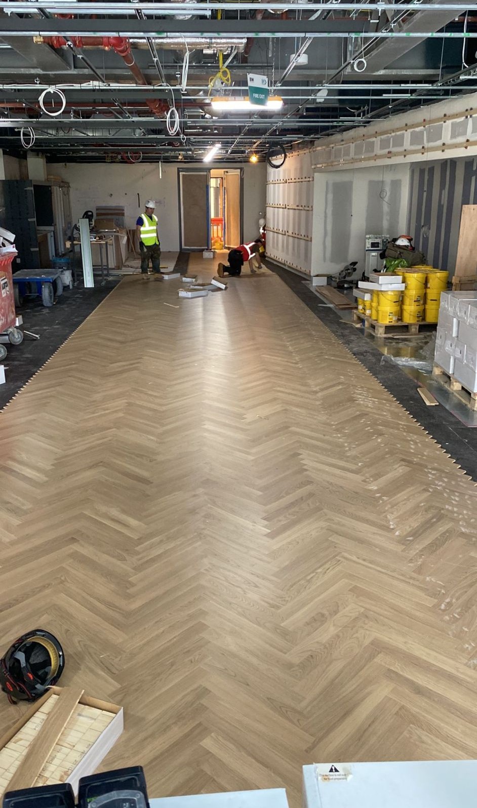 Large-scale herringbone timber parquet flooring being installed by flooring specialists inside a commercial interior under construction.