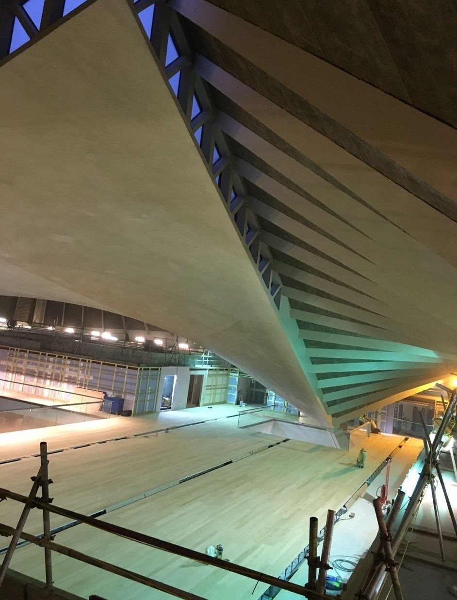 Expansive pale timber flooring beneath a dramatic ribbed architectural roof structure during construction and finishing works.