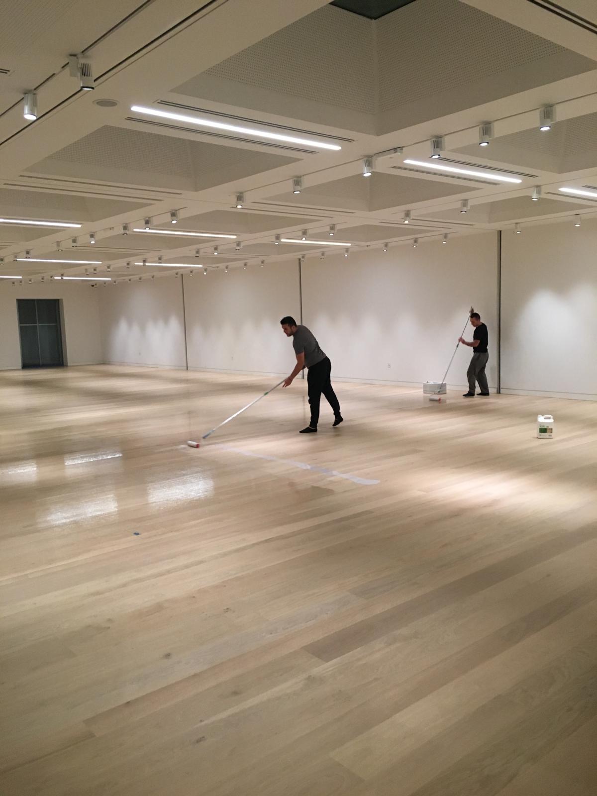 Two workers applying a finish to a pale timber floor in a large open room with ceiling lights.