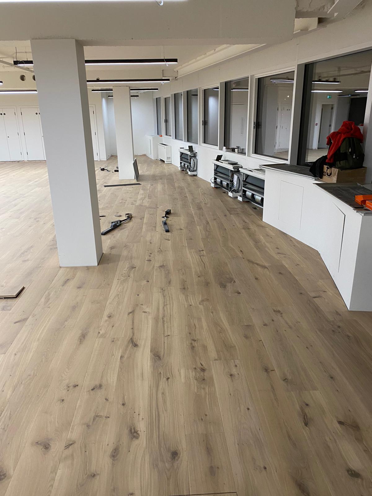 Wide natural timber plank flooring in a spacious contemporary interior with white columns and glass-partitioned rooms during installation works.