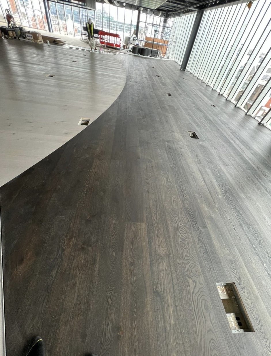 Dark timber flooring with a sweeping curved transition beside full-height windows during interior fit-out.